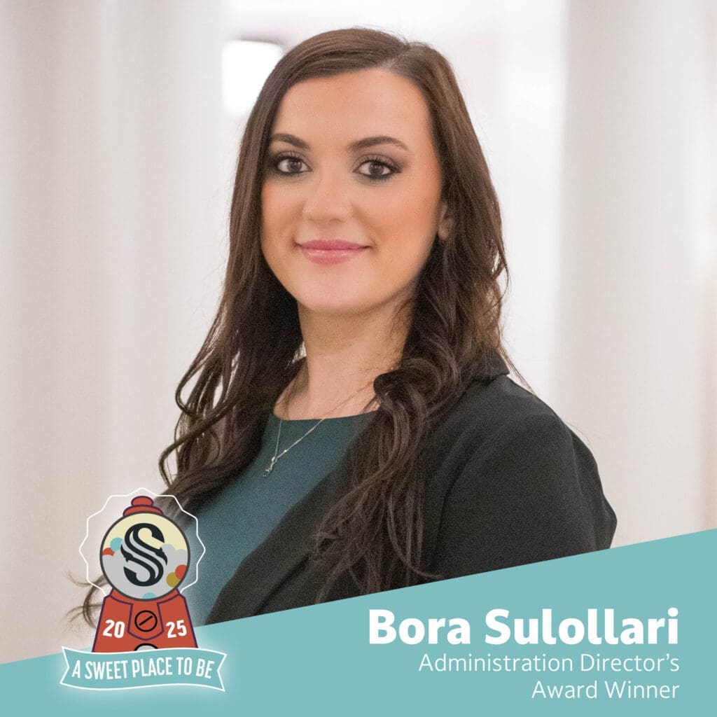 A woman with long brown hair, wearing a black blazer and green top, stands smiling in a bright hallway. Text reads "Bora Sulollari, Administration Director’s Award Winner." A badge says "2025, A Sweet Place to Be.