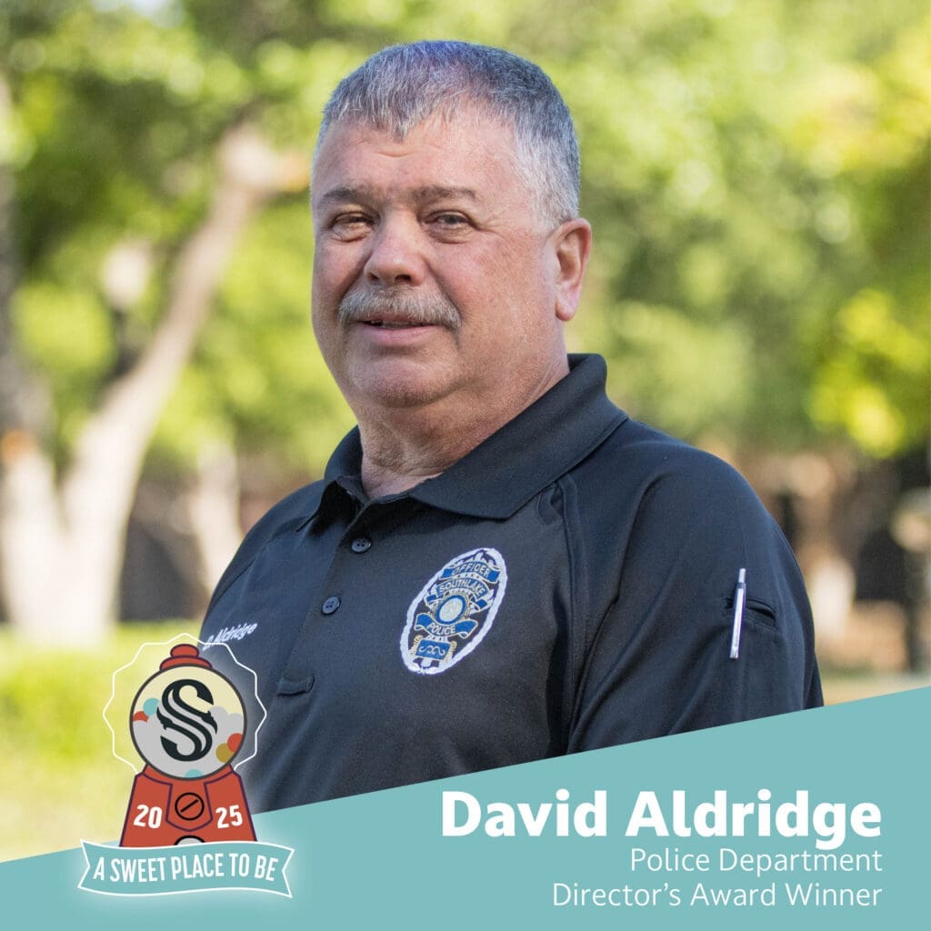 A man in a police uniform stands outdoors, smiling slightly. Text reads: "David Aldridge, Police Department, Director’s Award Winner." A badge graphic says "A Sweet Place to Be, 2025." Green trees are blurred in the background.