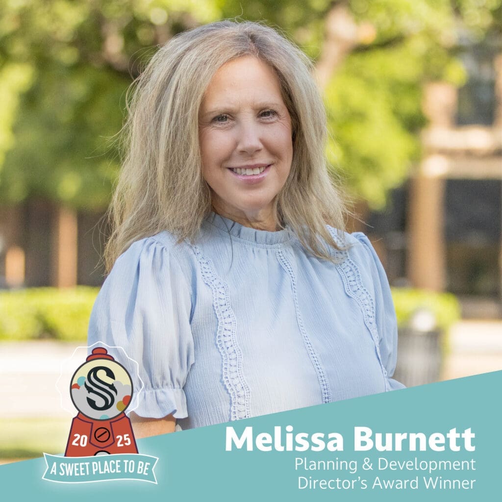 A woman with long blonde hair wearing a light blue blouse smiles outdoors. Text reads: “Melissa Burnett, Planning & Development Director’s Award Winner.” A logo says “2025 A Sweet Place To Be.” Green trees are blurred in the background.