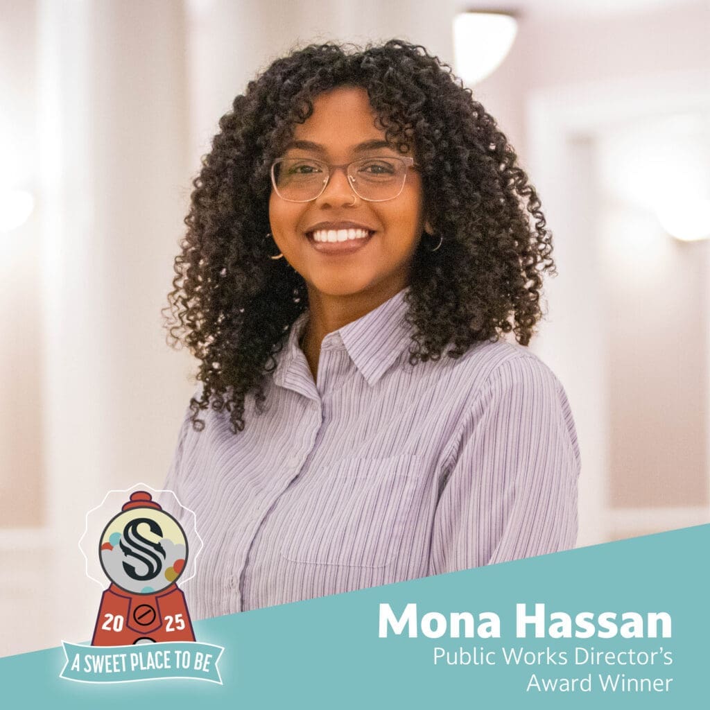 A woman with curly hair and glasses, wearing a striped button-up shirt, stands smiling indoors. Text reads: "Mona Hassan, Public Works Director’s Award Winner." A badge says "2025 A Sweet Place to Be.