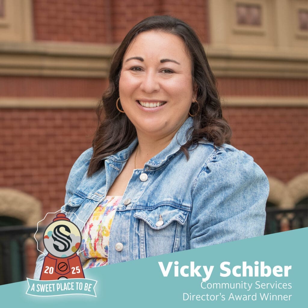A smiling woman with long dark hair wears a denim jacket over a floral top, standing in front of a brick building. Text reads: “Vicky Schiber, Community Services Director’s Award Winner.” A badge graphic is at the bottom left.