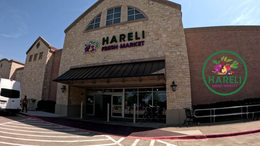 A stone storefront of Hareli Fresh Market with large green and purple signage above the entrance and a matching logo painted on the right wall. A white van and a shopping cart are visible outside.