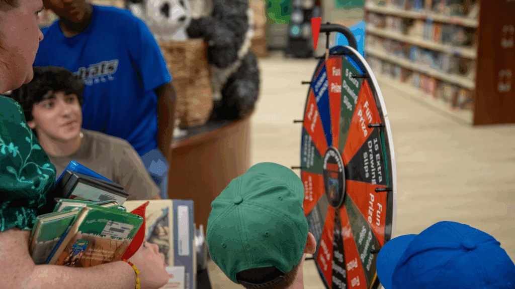 Several children, some wearing green and blue caps, gather around a colorful spinning prize wheel in a library. An adult holds a stack of books, and bookshelves and stuffed animals are visible in the background.