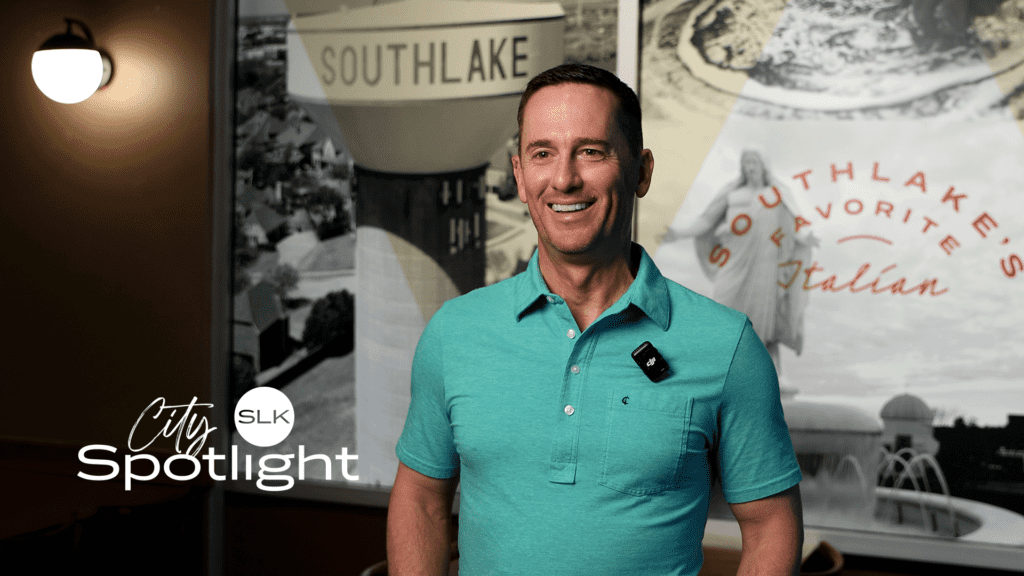 A smiling man in a teal polo stands indoors with a microphone clipped to his shirt. Behind him are images and text referencing Southlake, including "Southlake's Favorite Italian." City Spotlight SLK logo is in the lower left.