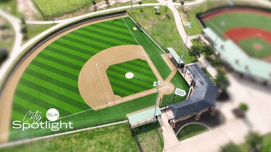 Aerial view of a well-maintained baseball field with green striped turf and tan infield, surrounded by pathways, trees, and nearby buildings. Text overlay reads “City Spotlight SLK.”.