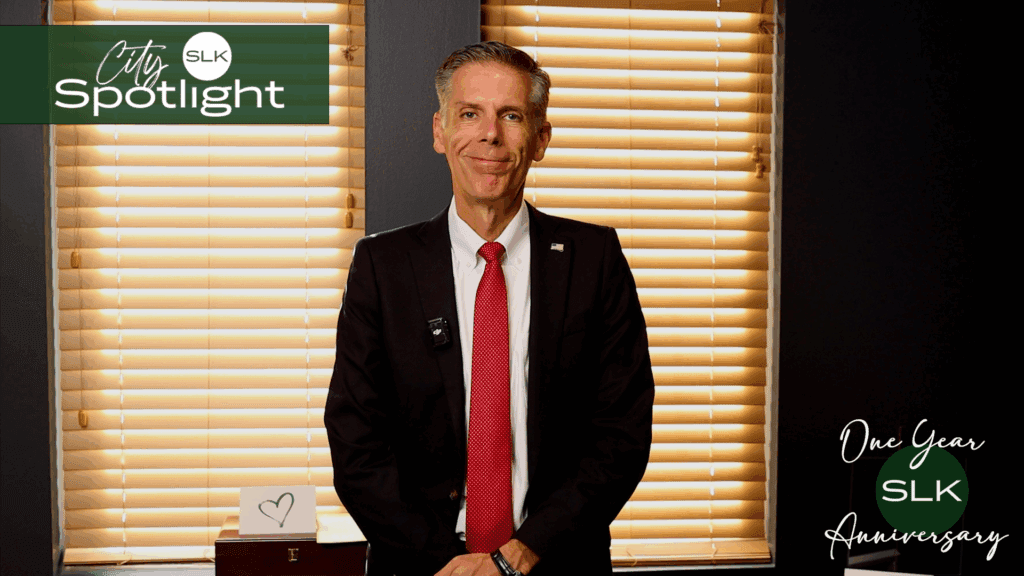 A man in a suit and red tie stands smiling in front of closed blinds. The image has City SLK Spotlight and One Year SLK Anniversary text, with a small heart card on a table in the background.