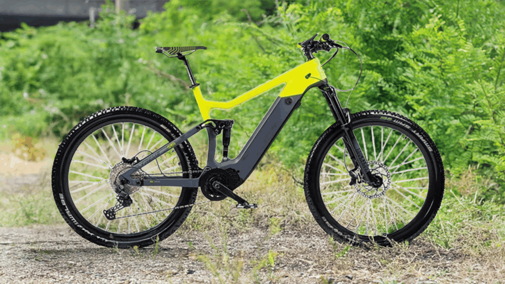 A modern electric mountain bike with a bright yellow and black frame stands on a gravel path surrounded by lush green bushes and grass.