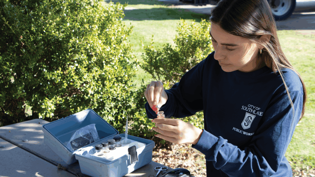 A woman wearing a "City of Southlake Public Works" shirt collects a water sample outdoors, using a dropper and small glass bottle next to a blue case with testing supplies on a table in a sunny park setting.