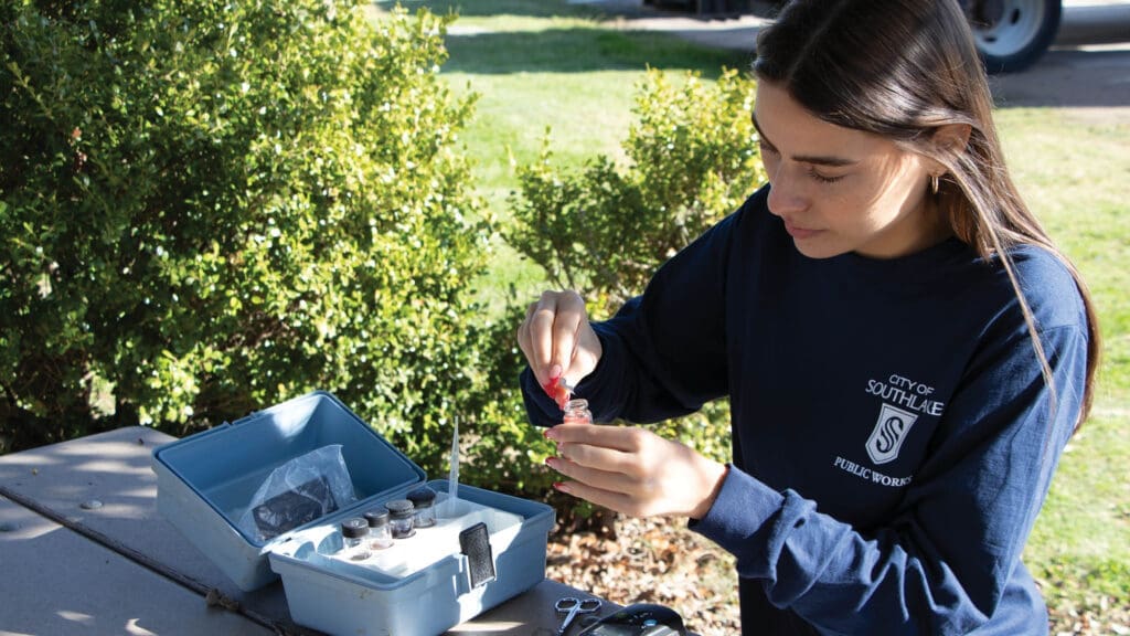 A woman in a "City of Southlake Public Works" shirt uses a dropper to add liquid to a small vial at an outdoor table, with testing supplies and greenery visible in the background.