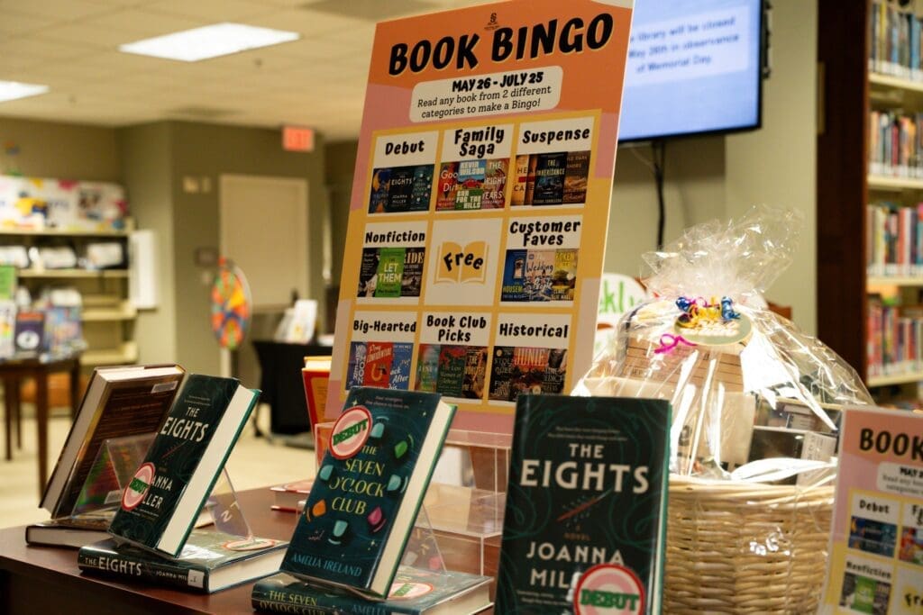 A library display features a Book Bingo sign with reading categories, several books on stands, and a raffle basket. Books include "The Eights" and "The Newcomer’s Book Club." Shelves of books are in the background.