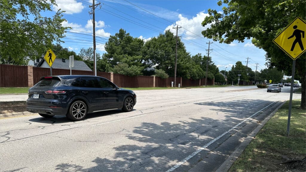 A dark SUV drives down a sunny suburban street with a yellow pedestrian crossing sign on the right. Trees, utility poles, and a wooden fence line the road under a blue sky with scattered clouds.