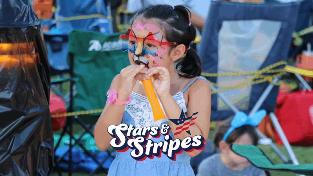 A young girl with colorful face paint eats an orange ice pop at an outdoor event. She wears red glasses and a blue dress. The words "Stars & Stripes" appear in front of her. Foldable chairs are in the background.