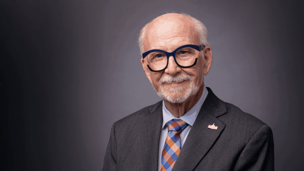An older man with glasses, a gray beard, and a mustache is wearing a suit, patterned tie, and a lapel pin, posing against a neutral gray background.