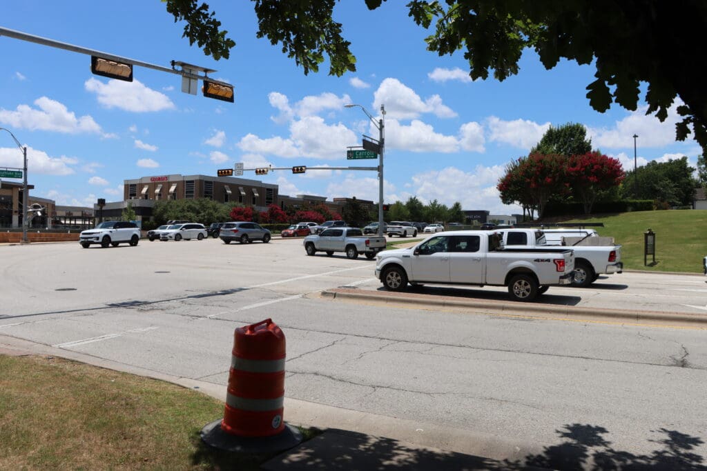 A busy intersection with white pickup trucks and other vehicles waiting at a traffic light, bordered by grassy areas and trees under a sunny, partly cloudy sky. A large orange traffic barrel sits on the sidewalk in the foreground.
