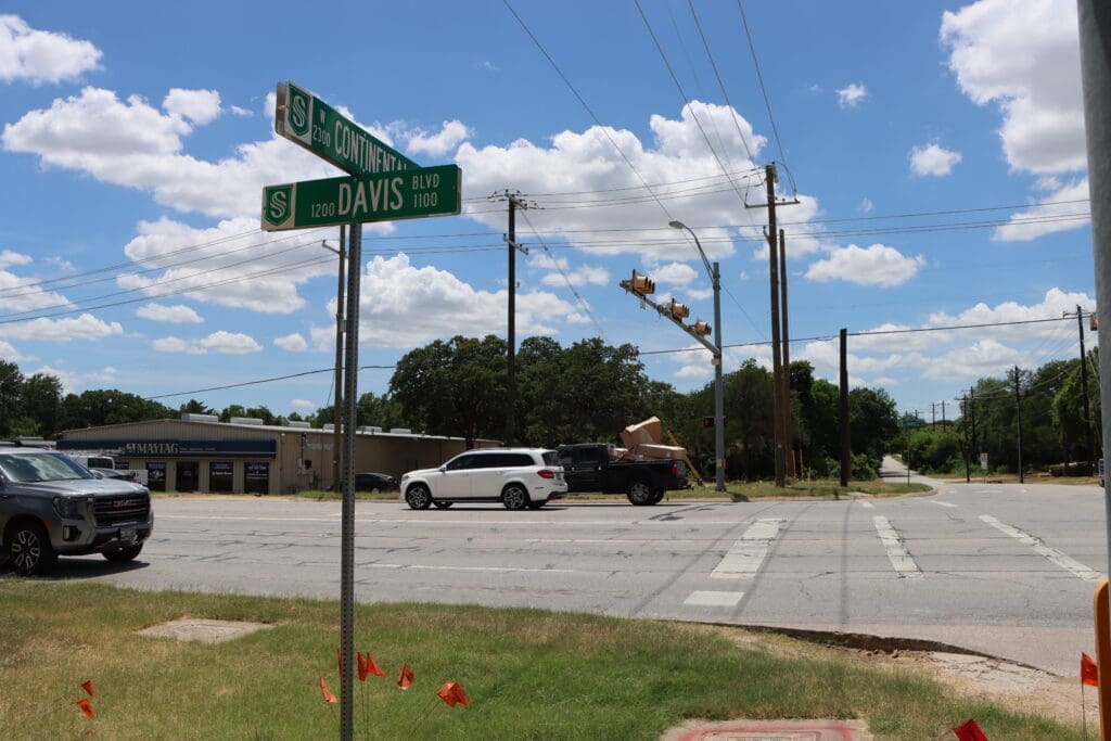 A street intersection with cars stopped at a traffic light under a blue sky. Street signs read "S. Davis Blvd" and "Continental." Trees and a building are in the background, with utility poles and traffic lights nearby.