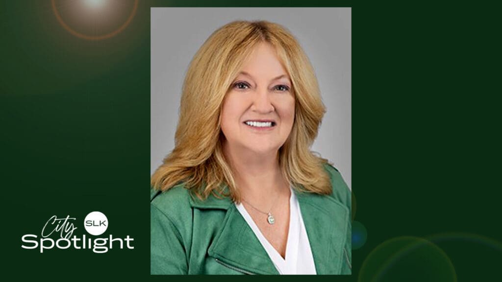 A woman with shoulder-length blonde hair, wearing a green jacket over a white top, smiling at the camera. The image has a dark green background with the text "City Spotlight SLK" in the corner.