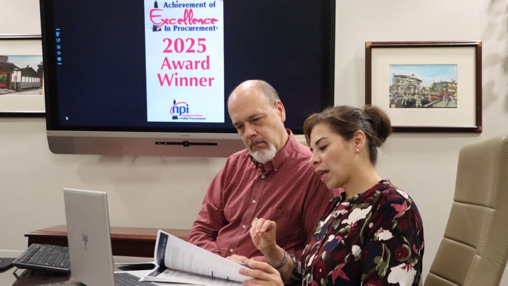 Two people sit at a desk reviewing a document together, with a laptop in front of them. Behind them, a screen displays a "2025 Award Winner" sign for excellence in procurement.