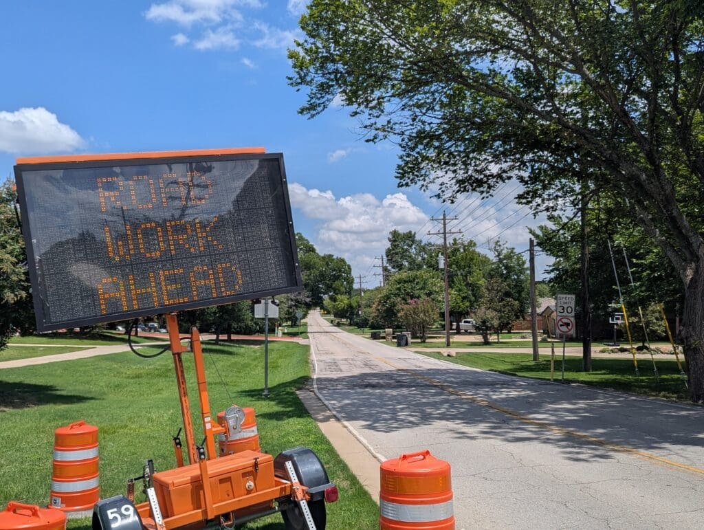 An electronic road sign displays "ROAD WORK AHEAD" next to orange traffic barrels on a quiet suburban street lined with trees under a blue sky with scattered clouds.