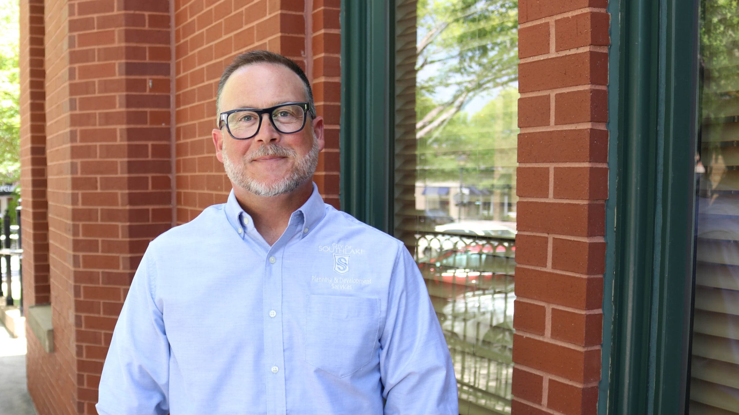 A man with glasses and a short beard, wearing a light blue button-up shirt, stands smiling in front of a red brick building with windows reflecting trees and parked cars.