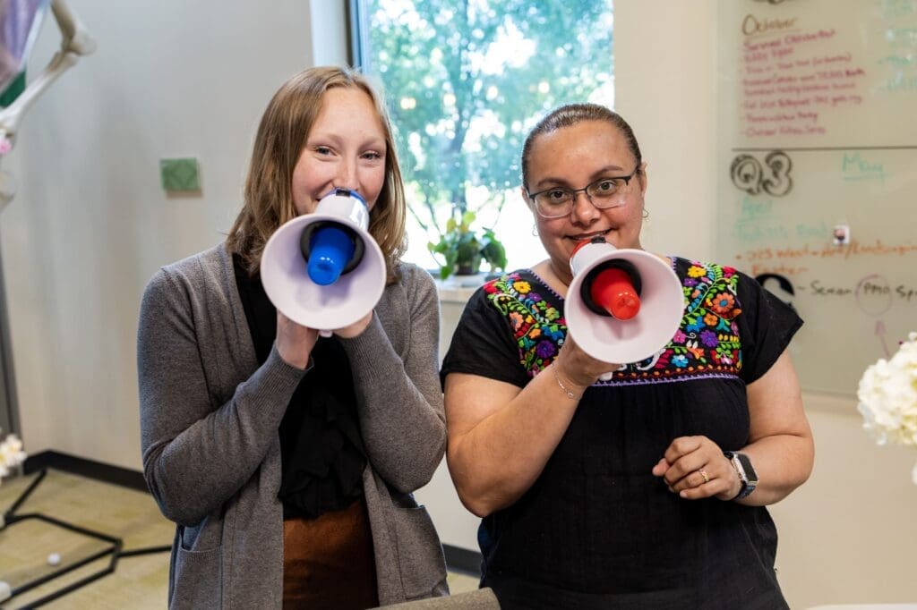 Two women standing indoors smile and speak into handheld megaphones. One wears a gray cardigan, the other glasses and a floral-embroidered shirt. A whiteboard and a window with trees are visible in the background.