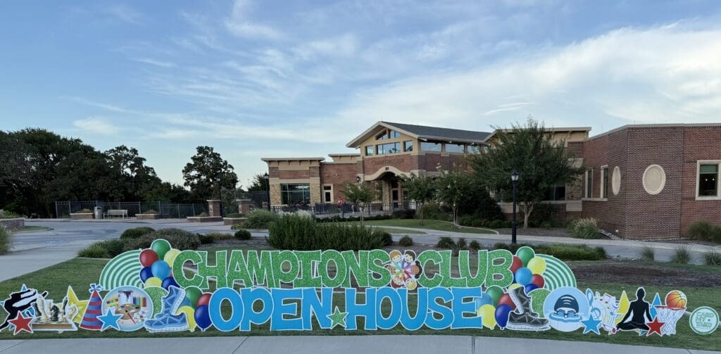A large colorful sign reading “Champions Club Open House” with balloons and various icons stands on the grass in front of a modern brick building under a blue sky.