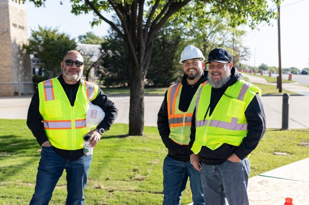 Three men wearing yellow safety vests and dark clothing stand outside on a sunny day. One wears a white hard hat, and they are smiling near a tree and grassy area, with buildings and a sidewalk in the background.