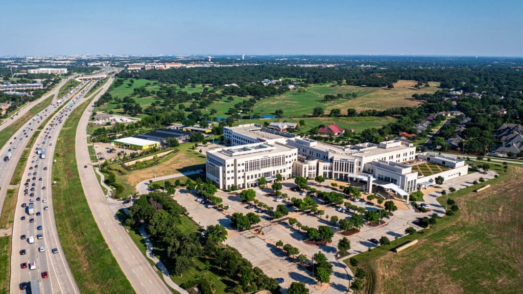 Aerial view of a large white building complex with a parking lot, adjacent to a busy freeway. Surrounding areas include green fields, trees, and suburban houses under a clear blue sky.