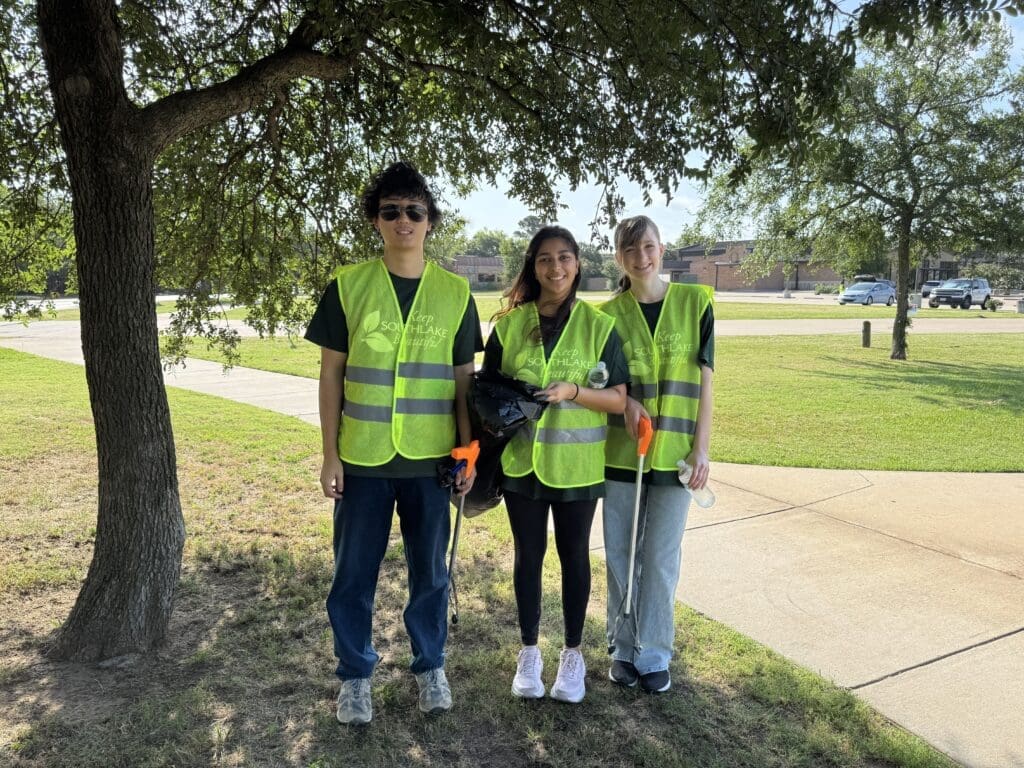 Three people wearing yellow safety vests stand under a tree on a grassy area near a sidewalk, smiling at the camera while holding trash grabbers and a black garbage bag.