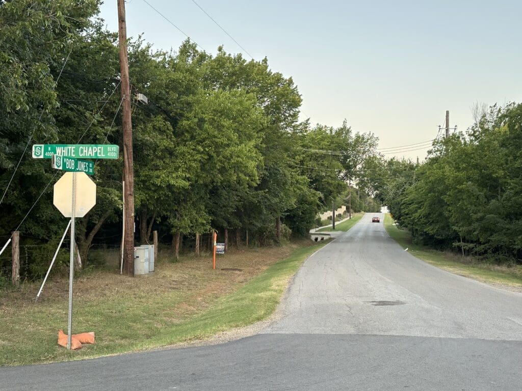 A quiet, empty road runs beside trees and utility poles. A street sign at the intersection reads "S WHITE CHAPEL RD" and "W BOB JONES ST." The sky is clear, and a single car is seen in the distance.