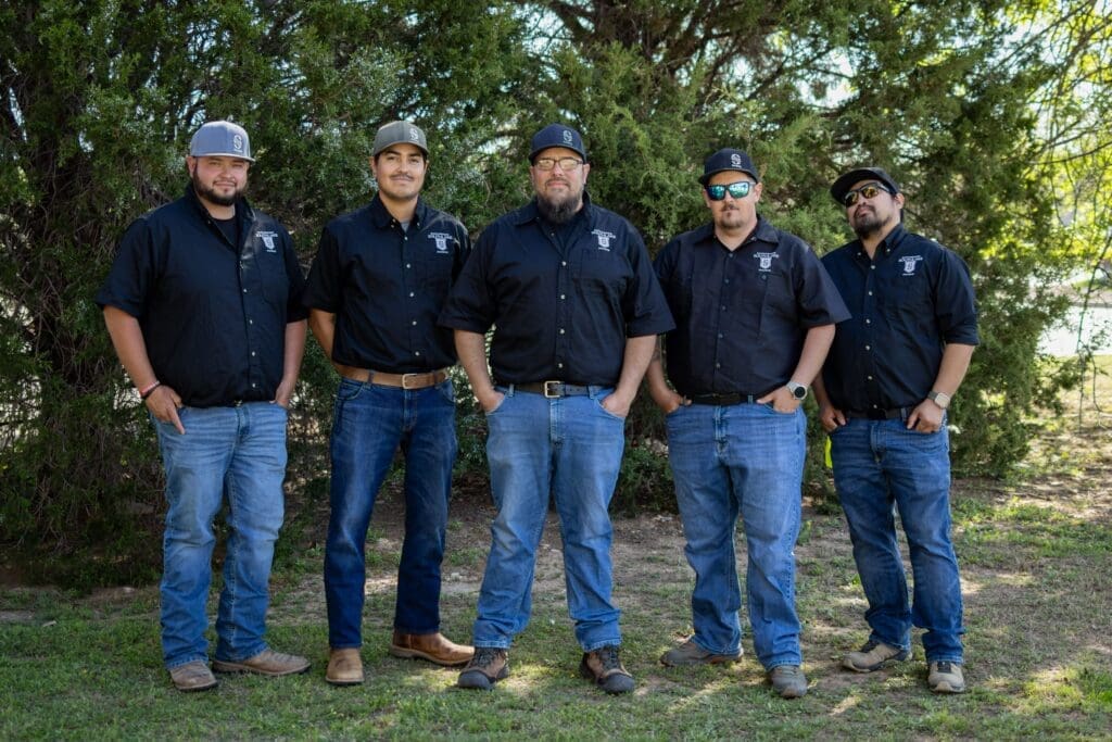 Five men stand outdoors in front of trees, all wearing black button-up shirts, blue jeans, and hats. They are smiling or posing casually with hands in pockets or arms relaxed.