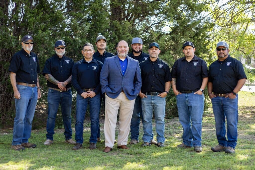 Nine men pose outdoors in front of trees; eight wear matching dark shirts, jeans, and caps, while one in the center wears a light suit and blue shirt. They all stand on grass, smiling at the camera.