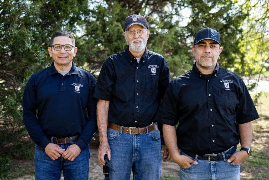 Three men stand outdoors in front of trees, all wearing black collared shirts with embroidered logos, blue jeans, and glasses or hats. They are smiling or looking at the camera in a sunny setting.