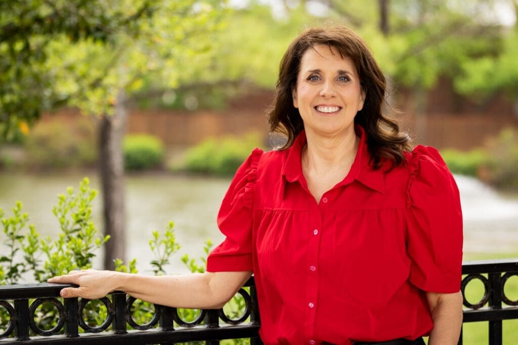 A woman with brown hair wearing a bright red blouse stands outdoors, smiling, with one hand resting on a black metal fence. Green trees and a body of water are visible in the blurred background.
