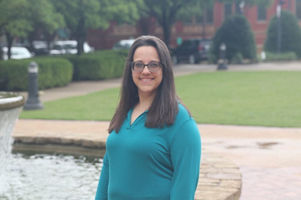 A woman with long brown hair and glasses, wearing a teal top, stands and smiles near a stone-edged fountain in a park with green lawns and trees in the background.