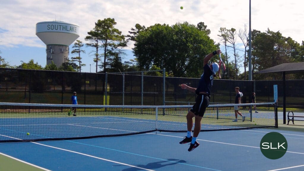 A tennis player jumps to hit a serve on an outdoor court. Other players and a coach stand nearby. Trees and a large water tower labeled "SOUTHLAKE" are visible in the background. The SLK logo appears in the corner.