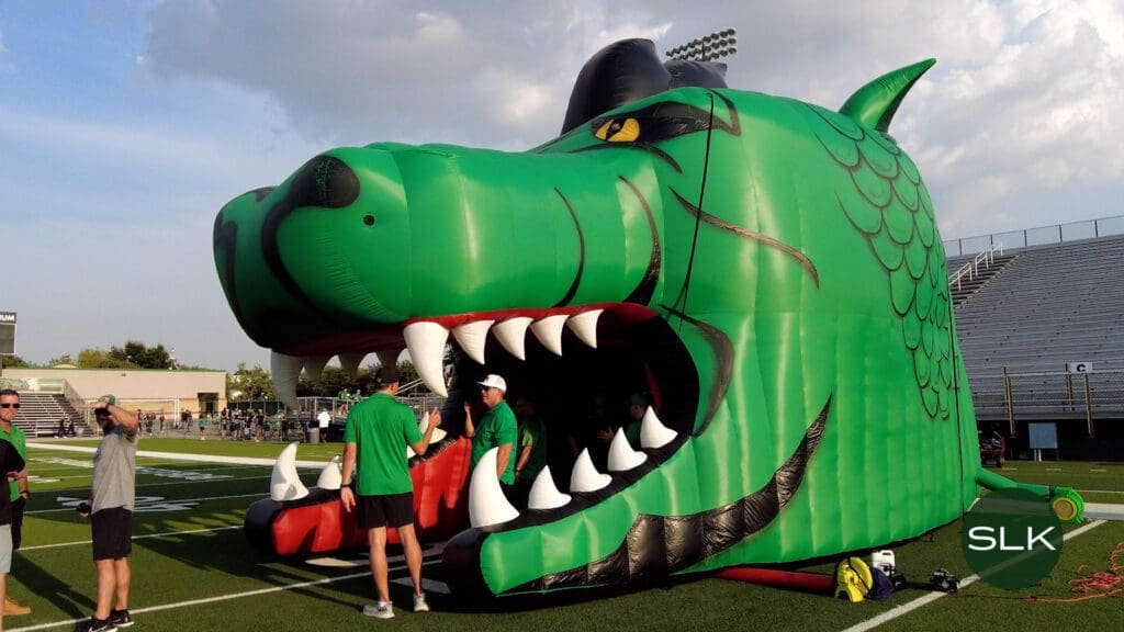 A large inflatable green dragon head with open mouth and white fangs sits on a football field, with several people in green shirts standing nearby. Bleachers and stadium lights are visible in the background. An icon with a green circle and "SLK" is in the bottom right corner.