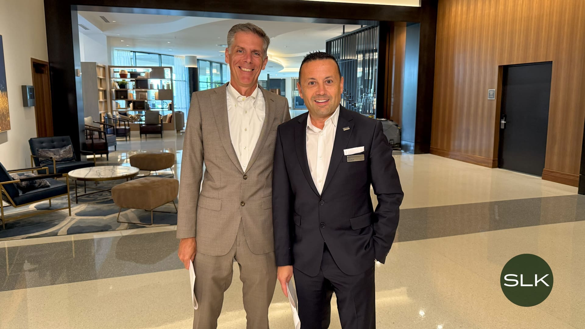 Two men in business suits stand smiling in a modern, well-lit hotel lobby. One holds papers, and both wear name tags. The space features elegant seating and wood accents. A circular SLK logo appears in the lower right corner.