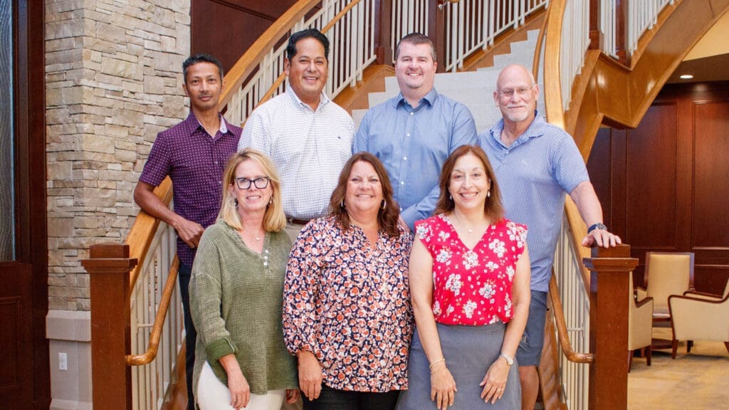 Seven adults pose and smile together on a staircase in a well-lit, modern interior with wooden railings and stonework. Four stand at the back and three are in front. The setting appears formal yet friendly.