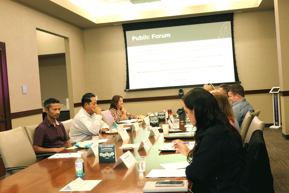 A group of people sit around a conference table in a meeting room, engaged in discussion. A projector displays a “Public Forum” slide on the screen at the front of the room. Nameplates and notepads are on the table.