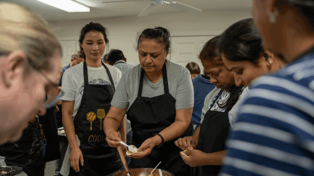 A group of people in aprons gather around a table, focused on preparing food together in a well-lit kitchen. One woman in the center carefully fills a dumpling wrapper.