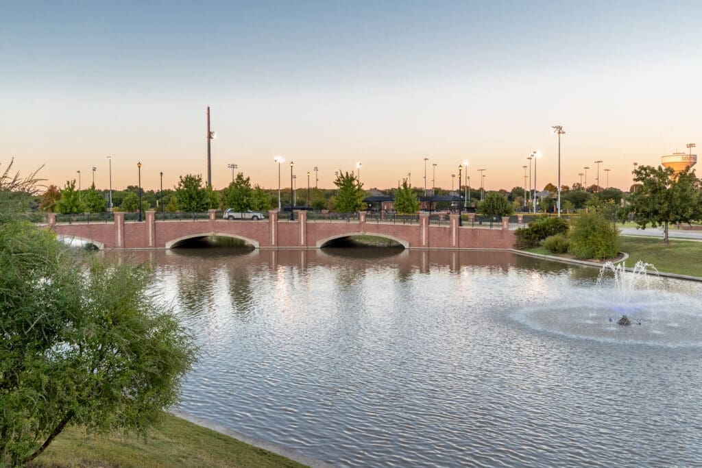 A brick bridge with three arches spans a calm pond, with a water fountain on the right. Trees and light poles line the background, under a clear sky at sunset.