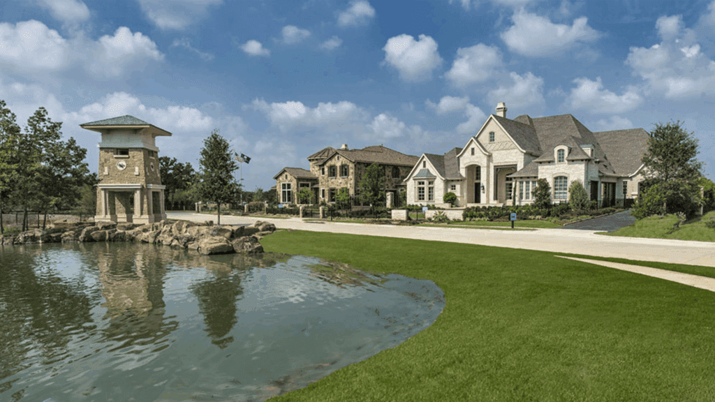 Large upscale houses with varied architecture sit behind a manicured lawn, next to a small pond with rocks and a stone tower structure under a blue sky with scattered clouds.