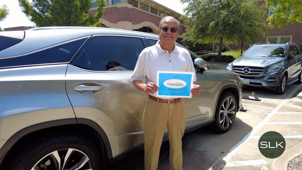 A smiling man in sunglasses and light clothing stands in front of a silver SUV, holding a sign that says "CALL A RIDE SOUTHLAKE" in a parking lot with trees and a building in the background.
