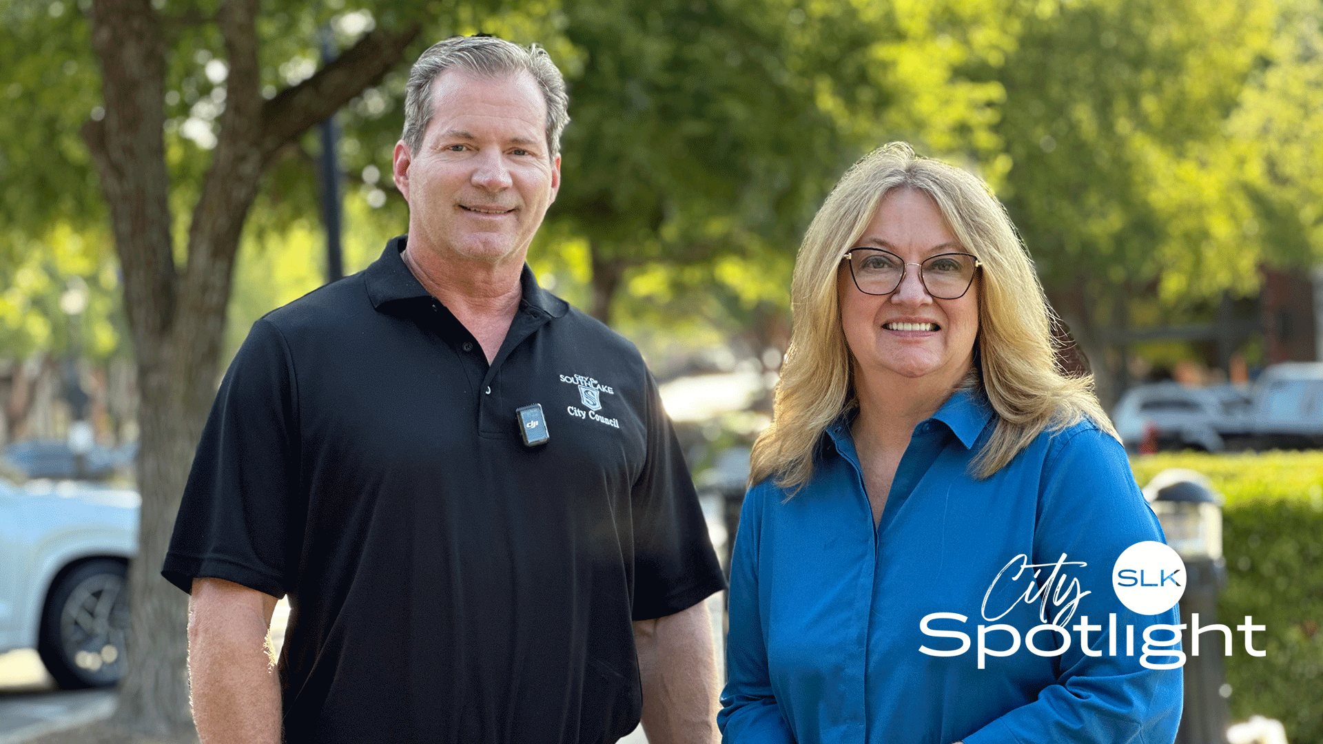 Councilmember Chuck Taggart and Deputy Mayor Pro Tem Kathy Talley smiling outdoors on a sunny day—one wearing a black polo shirt and the other a blue blouse. Green trees and parked cars are in the background. The words "City SLK Spotlight" appear in the corner.