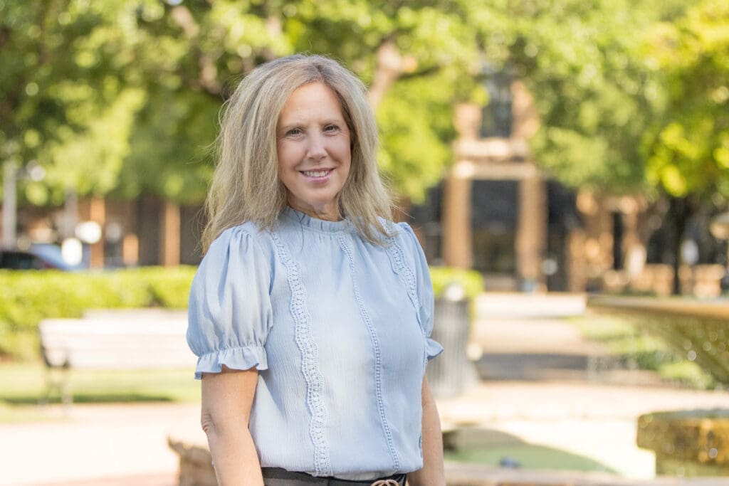 A woman with long blonde hair wearing a light blue blouse stands outdoors in a sunny park, smiling at the camera. Green trees and a bench are visible in the blurred background.