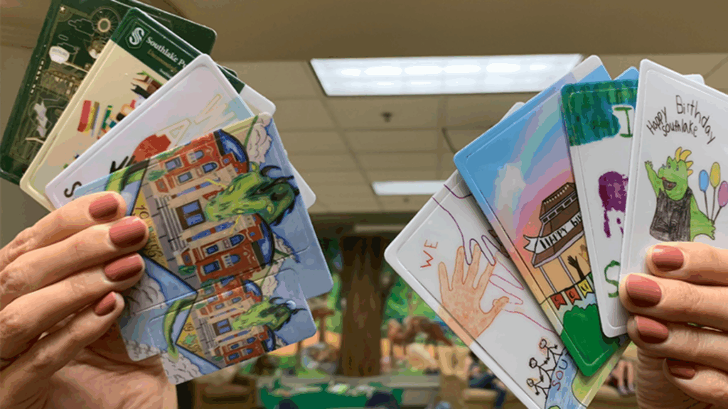 Two hands with painted nails hold several colorful illustrated cards featuring buildings, birthday themes, hands, and cartoon green dragons, with a blurred indoor background.