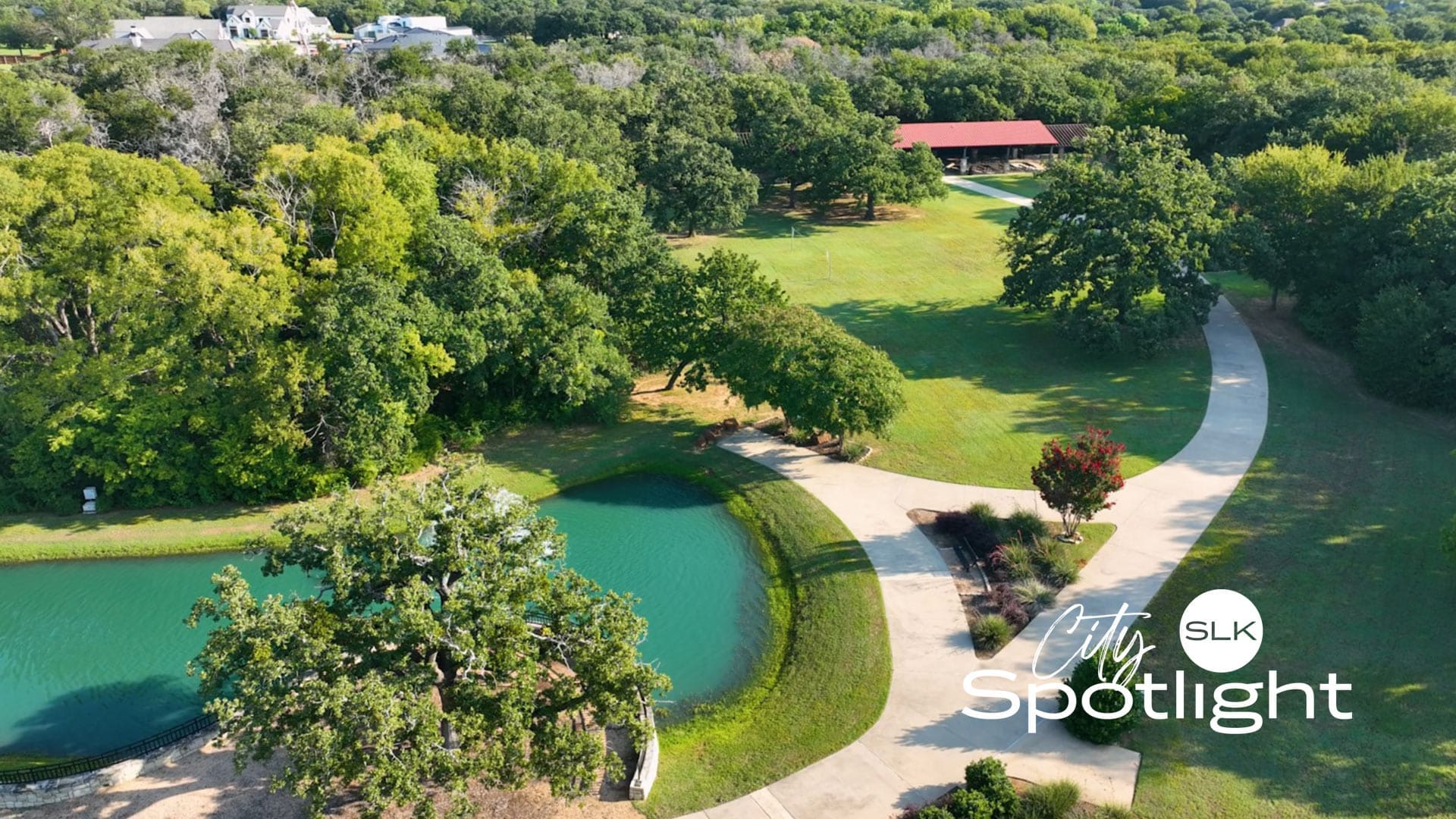 Aerial view of a green park with a pond, winding pathways, trees, and a large pavilion. The text "City Spotlight SLK" is overlaid on the lower right corner.