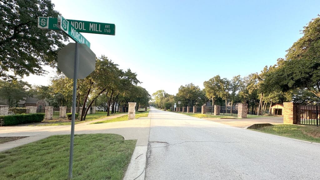 A wide suburban street lined with trees and brick pillars. A street sign at the corner reads "S Pool Mill Ave" and "Hackamore Dr." The sky is clear and the area is calm and quiet.