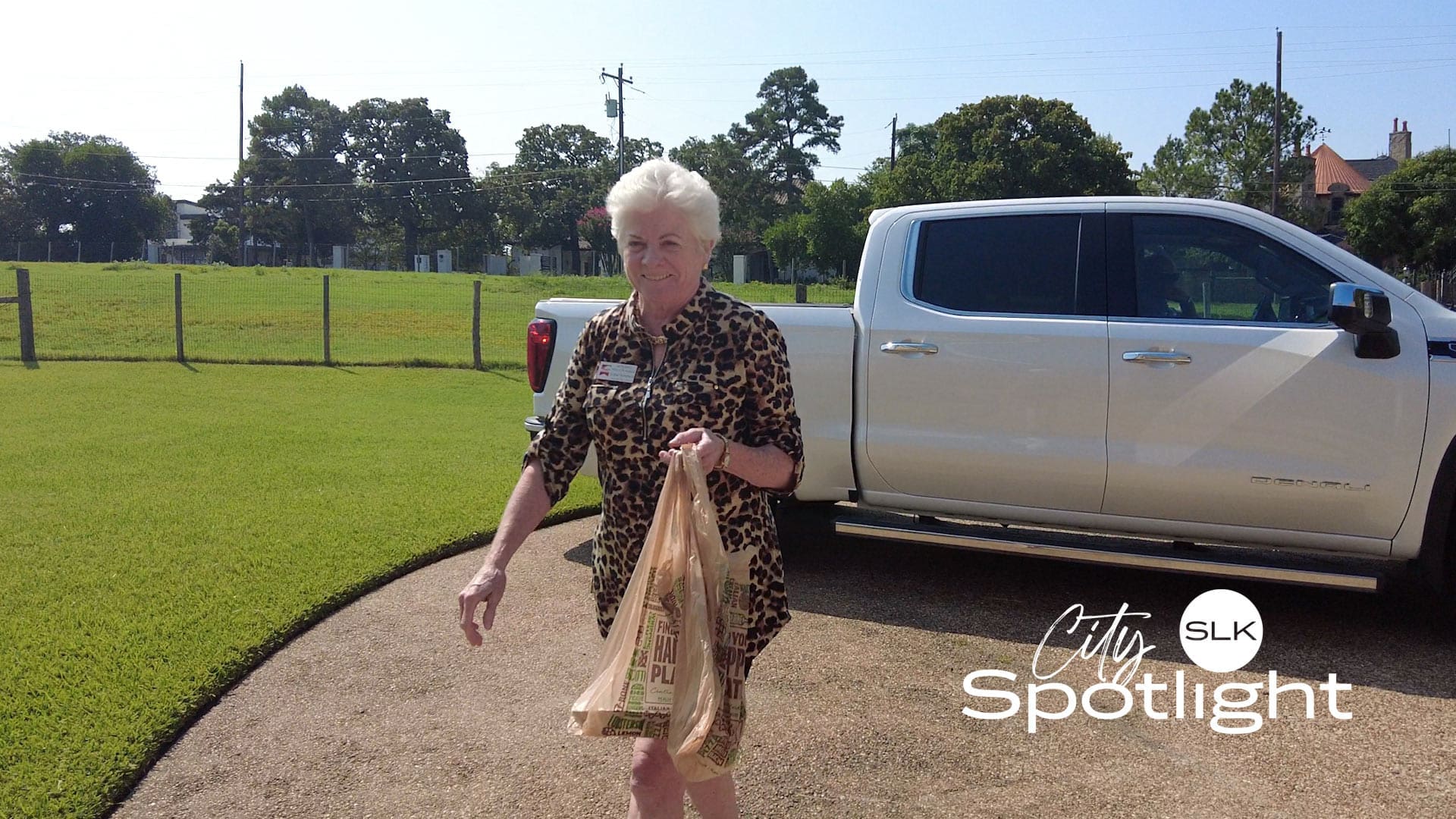 An older woman in a leopard print dress carries groceries while walking on a driveway near a white pickup truck. It is sunny, with green grass and trees in the background. The words "City Spotlight SLK" are overlaid on the image.