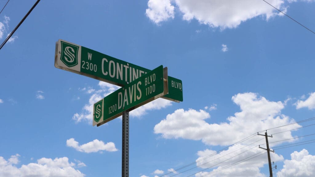 A street sign at the intersection of Continental Blvd (2300 W) and Davis Blvd (1200 S, 1100 S) against a blue sky with scattered clouds. Power lines are visible in the background.